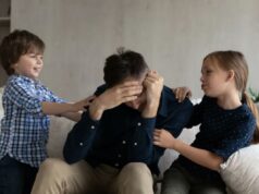 Papai foi criticado por se sentir ‘destruído’ por estar preso em casa com os filhos: ‘Infuriantemente exaustivo’ Um pai exausto está sentado em um sofá com o rosto entre as mãos, enquanto seus dois filhos pequenos, um menino e uma menina, ficam um de cada lado, tocando-o.