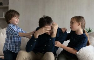 Papai foi criticado por se sentir ‘destruído’ por estar preso em casa com os filhos: ‘Infuriantemente exaustivo’ Um pai exausto está sentado em um sofá com o rosto entre as mãos, enquanto seus dois filhos pequenos, um menino e uma menina, ficam um de cada lado, tocando-o.