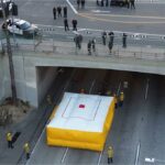 Vista aérea de uma passagem subterrânea de uma rodovia com amortecedores amarelos e caminhões de bombeiros, indicando um incidente.