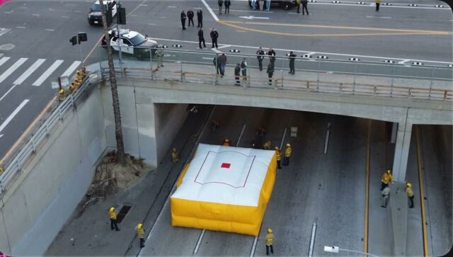 Vista aérea de uma passagem subterrânea de uma rodovia com amortecedores amarelos e caminhões de bombeiros, indicando um incidente.
