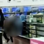 Um policial de uniforme escuro fica de costas para a câmera dentro de uma loja, com clientes e mercadorias visíveis ao seu redor.