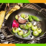 Duas panelas de aço carbono com frango assado e brócolis, e batatas assadas, na grelha a carvão.