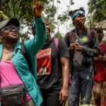 Uma jovem com um chapéu largo segura um peixe perto de um rio, sorrindo
