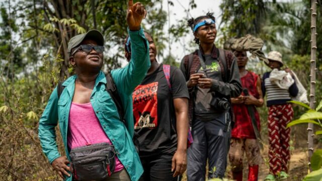 Uma jovem com um chapéu largo segura um peixe perto de um rio, sorrindo 