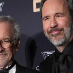 Um homem mais velho, de cabelos grisalhos e barba, fala ao microfone, vestindo um paletó esporte, diante de um fundo cinza suave.