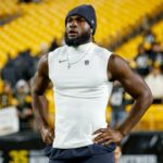 Um jogador de futebol americano vestindo um gorro e uma camisa sem mangas com o logotipo da NFL, com as mãos na cintura, em pé no campo com assentos amarelos ao fundo.