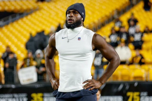 Um jogador de futebol americano vestindo um gorro e uma camisa sem mangas com o logotipo da NFL, com as mãos na cintura, em pé no campo com assentos amarelos ao fundo.