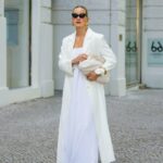 Diane Batoukina usa vestido midi de seda branca com estampa de listras azuis e marinho, mangas curtas e gola pontuda da Sandro. Um lenço de seda Sandro combinando está amarrado frouxamente no decote. Uma mini bolsa branca com alça de corrente e detalhes dourados da Chanel é usada na transversal. O calçado consiste em sandálias brancas de salto aberto da Aeyde. Os óculos de sol quadrados Dior com lentes gradientes escuras completam o visual. Cabelo castanho longo e liso é penteado com uma parte central. Os lábios ficam sutilmente coloridos e a pele parece lisa e uniforme. Foto de corpo inteiro, durante uma sessão de fotos de moda de rua, em 28 de abril de 2025 em Paris, França.