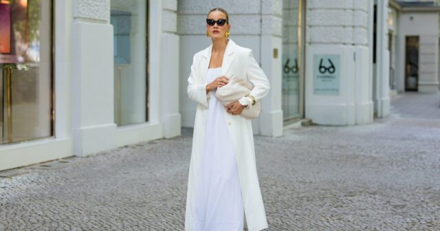 Diane Batoukina usa vestido midi de seda branca com estampa de listras azuis e marinho, mangas curtas e gola pontuda da Sandro. Um lenço de seda Sandro combinando está amarrado frouxamente no decote. Uma mini bolsa branca com alça de corrente e detalhes dourados da Chanel é usada na transversal. O calçado consiste em sandálias brancas de salto aberto da Aeyde. Os óculos de sol quadrados Dior com lentes gradientes escuras completam o visual. Cabelo castanho longo e liso é penteado com uma parte central. Os lábios ficam sutilmente coloridos e a pele parece lisa e uniforme. Foto de corpo inteiro, durante uma sessão de fotos de moda de rua, em 28 de abril de 2025 em Paris, França.