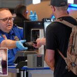 Um agente da TSA com uniforme azul e luvas devolve um passaporte a um viajante com uma mochila.