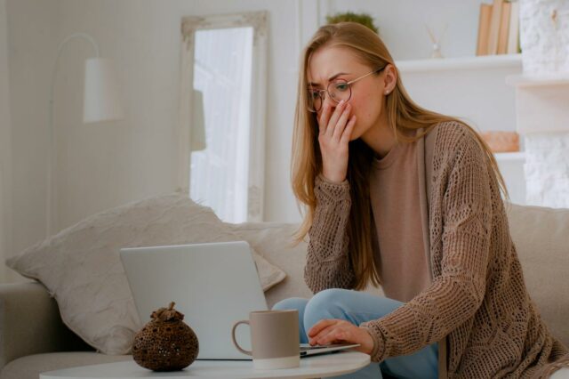 Mulher de óculos parecendo chocada com a tela de um laptop, cobrindo a boca com a mão.