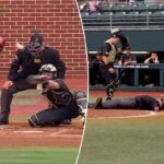 Um árbitro de beisebol está deitado de bruços no chão, atrás do home plate, com um receptor próximo e outros jogadores no banco de reservas.