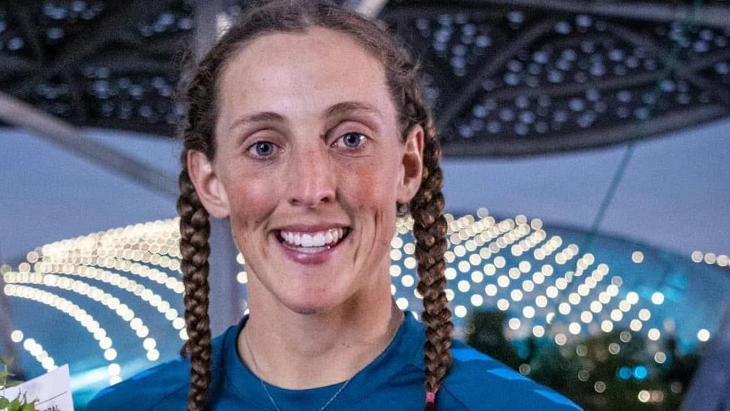 Uma mulher com kit de esportes aquáticos, segurando uma canoa e um remo. Ela tem cabelos castanhos presos em duas tranças e está sorrindo e olhando para a câmera. Atrás dela há um quadro com vários logotipos de empresas.