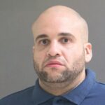 Mugshot de um homem careca com barba vestindo uma camisa pólo azul.
