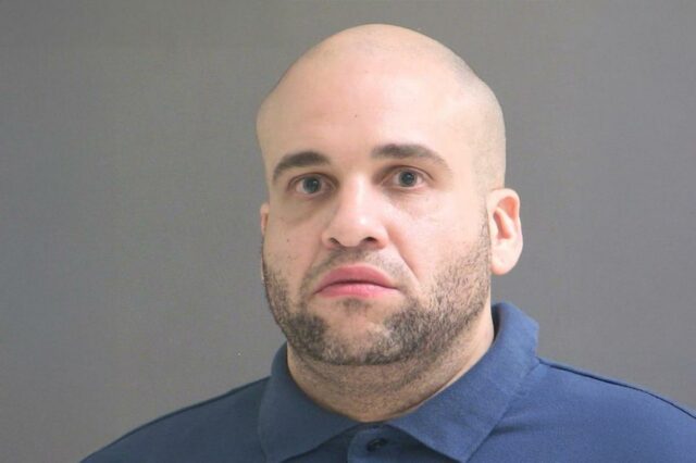 Mugshot de um homem careca com barba vestindo uma camisa pólo azul.
