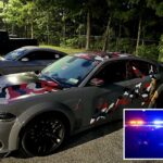 Dois muscle cars, um com camuflagem vermelha, branca e cinza, estacionados no asfalto.