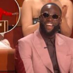 Draymond Green ri enquanto usa óculos escuros redondos e um terno rosa sobre uma camisa escura e gravata, sentado em uma cadeira vermelha.