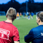 Treinadores de futebol assistindo a um treino noturno no campo de futebol de base durante o programa comunitário de desenvolvimento do futebol. Entain pressiona regulador do futebol visando patrocínios de jogos de azar ilegais em clubes da Premier League