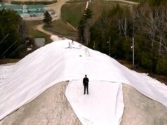 Esta colina de esqui quer guardar neve para a próxima temporada. Isso pode ser feito? Uma foto de drone mostra uma enorme pilha de neve, camadas de lonas e um homem em pé no topo da pilha.