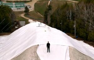 Esta colina de esqui quer guardar neve para a próxima temporada. Isso pode ser feito? Uma foto de drone mostra uma enorme pilha de neve, camadas de lonas e um homem em pé no topo da pilha.
