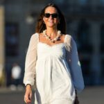 Diane Batoukina usa vestido midi de seda branca com estampa de listras azuis e marinho, mangas curtas e gola pontuda da Sandro. Um lenço de seda Sandro combinando está amarrado frouxamente no decote. Uma mini bolsa branca com alça de corrente e detalhes dourados da Chanel é usada na transversal. O calçado consiste em sandálias brancas de salto aberto da Aeyde. Os óculos de sol quadrados Dior com lentes gradientes escuras completam o visual. Cabelo castanho longo e liso é penteado com uma parte central. Os lábios ficam sutilmente coloridos e a pele parece lisa e uniforme. Foto de corpo inteiro, durante uma sessão de fotos de moda de rua, em 28 de abril de 2025 em Paris, França.