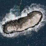 Vista aérea de uma pequena ilha rochosa cercada por ondas em um mar azul escuro.