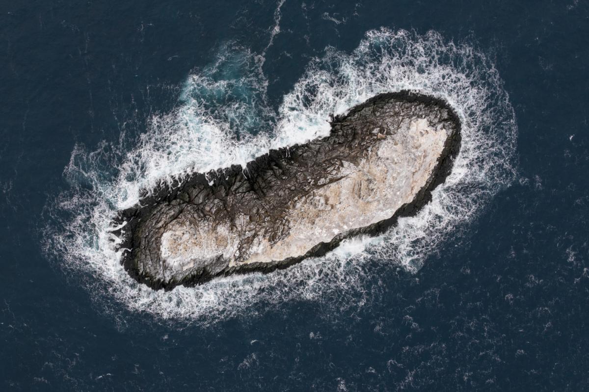 Vista aérea de uma pequena ilha rochosa cercada por ondas em um mar azul escuro.