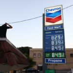 Um homem parado em um telhado em frente a uma placa de posto de gasolina da Chevron que exibe preços de gasolina acima de US$ 8,00 por galão.