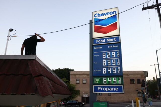 Um homem parado em um telhado em frente a uma placa de posto de gasolina da Chevron que exibe preços de gasolina acima de US$ 8,00 por galão.