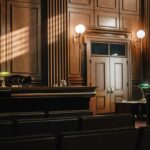 Ilustração de um tribunal vazio em estilo americano, com painéis e assentos de madeira, com banco de juiz, mesas para defesa e renomados e grandes janelas.