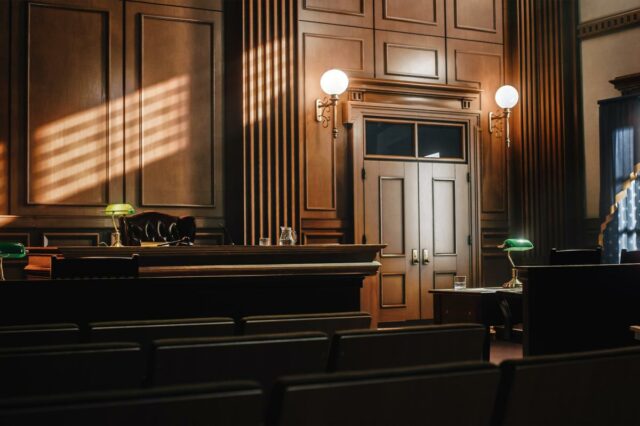 Ilustração de um tribunal vazio em estilo americano, com painéis e assentos de madeira, com banco de juiz, mesas para defesa e renomados e grandes janelas.