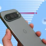 Uma mão segurando um smartphone Google Pixel azul com um ícone de alerta e ícones representando câmera, áudio e bugs.