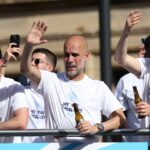 O técnico cessante Pep Guardiola acena enquanto os jogadores posam ao lado de todos os troféus conquistados ao longo da carreira de Pep Guardiola no City em um evento pós-festa com os jogadores após suas vitórias nas duas competições da copa nacional em 25 de maio de 2026, no centro de Manchester, noroeste da Inglaterra. (Foto de Darren Staples/AFP)