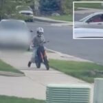 Um jovem em uma bicicleta suja vermelha com um capacete branco dirige em uma calçada, com um carro cinza embaçado atrás dele.