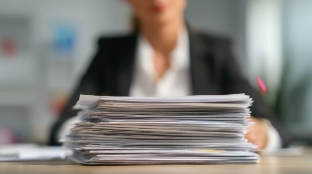 Novo na contratação de funcionários? Veja como revisar currículos sem Uma pessoa revisando um currículo, com outros currículos, material de escritório e uma lupa em uma mesa de madeira.