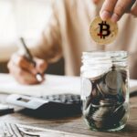 Uma pessoa segura uma moeda Bitcoin enquanto olha para um aplicativo de negociação de criptomoedas em um smartphone, com tabelas e gráficos em uma tela desfocada ao fundo.