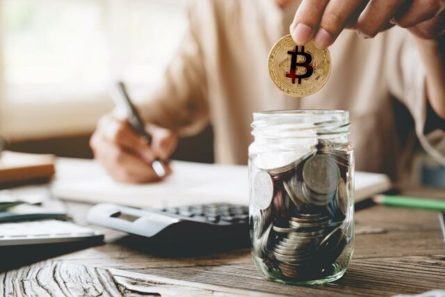 O Bitcoin é um bom investimento neste momento? O que Uma pessoa segura uma moeda Bitcoin enquanto olha para um aplicativo de negociação de criptomoedas em um smartphone, com tabelas e gráficos em uma tela desfocada ao fundo.