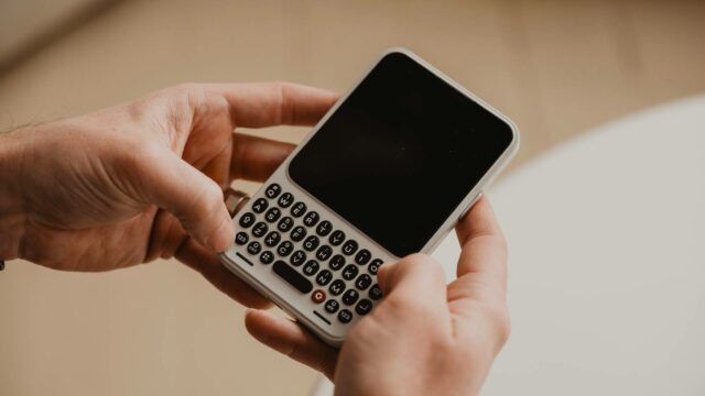 O Clicks Communicator está recebendo um grande aumento de bateria Uma mão segurando o Comunicador Clicks, flanqueado por vistas angulares destacadas.