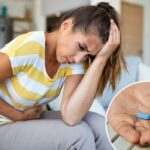 Mulher sentada em um sofá, segurando a cabeça e o estômago com dor, sofrendo de enxaqueca e cólicas abdominais.