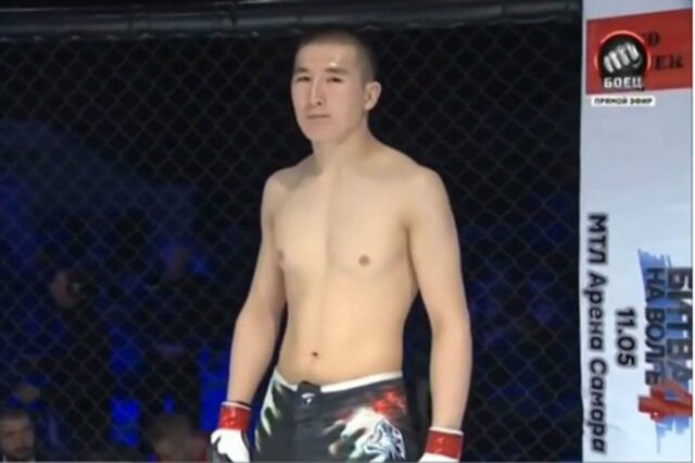 Medet Zheenaliev, sem camisa e usando shorts e luvas de luta, em uma jaula de combate.