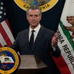 Gavin Newsom falando em um pódio com as bandeiras dos EUA e da Califórnia atrás dele.