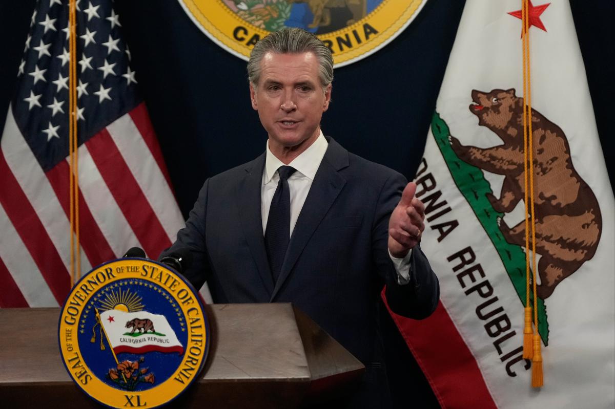 Gavin Newsom falando em um pódio com as bandeiras dos EUA e da Califórnia atrás dele.