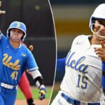 Uma jogadora de softball da UCLA, uniformizada com capacete azul e protetor de queixo branco, levanta a mão direita e aponta.