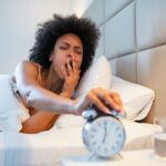Uma jovem negra sonolenta com um cabelo afro boceja enquanto pega a soneca em um despertador prateado.