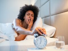 Os cientistas encontraram o ponto ideal para dormir – durma menos ou mais e você envelhecerá mais rápido Uma jovem negra sonolenta com um cabelo afro boceja enquanto pega a soneca em um despertador prateado.