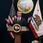 O governador Gavin Newsom fala em um pódio próximo a uma tela que exibe estatísticas econômicas dos EUA e imagens de Joe Biden e Donald Trump.