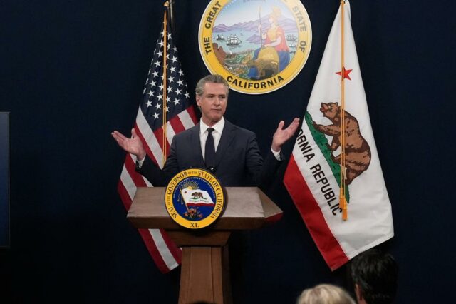 O governador Gavin Newsom fala em um pódio próximo a uma tela que exibe estatísticas econômicas dos EUA e imagens de Joe Biden e Donald Trump.