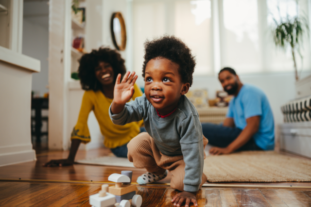 A stock photo showing a child playing a game with a train toy while his parents are laughing from the background.