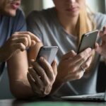 Um homem e uma mulher usam smartphones enquanto um laptop está aberto na mesa entre eles.