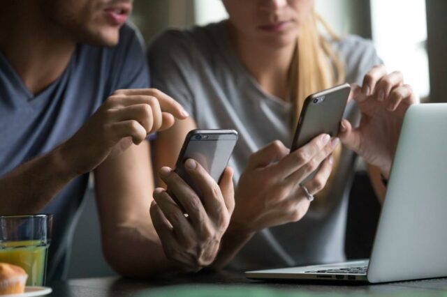 Os titãs da tecnologia podem ganhar mais de US$ 1 Um homem e uma mulher usam smartphones enquanto um laptop está aberto na mesa entre eles.